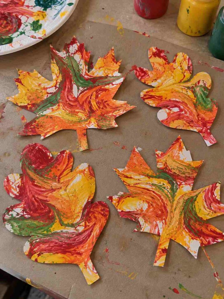 21 Easy & Creative Fall Crafts for Kids to Make at Home 11 9. Marble Fall Leaves Craft for Kids
Create colorful autumn leaves with simple paint swirls. Kids can mix fall colors, then cut the painted paper into leaf shapes for a fun, easy fall craft.
Materials Needed:
- White paper
- Red, orange, yellow, and green paint
- Spoon or craft stick
- Pencil and child-safe scissors
How to Make It:
1. Add small drops of paint to white paper.
2. Gently swirl the colors with a spoon or craft stick.
3. Let the paper dry completely.
4. Draw leaf shapes and cut them out.
Quick Tip: Don’t mix the colors too much—pretty swirls make every leaf unique.